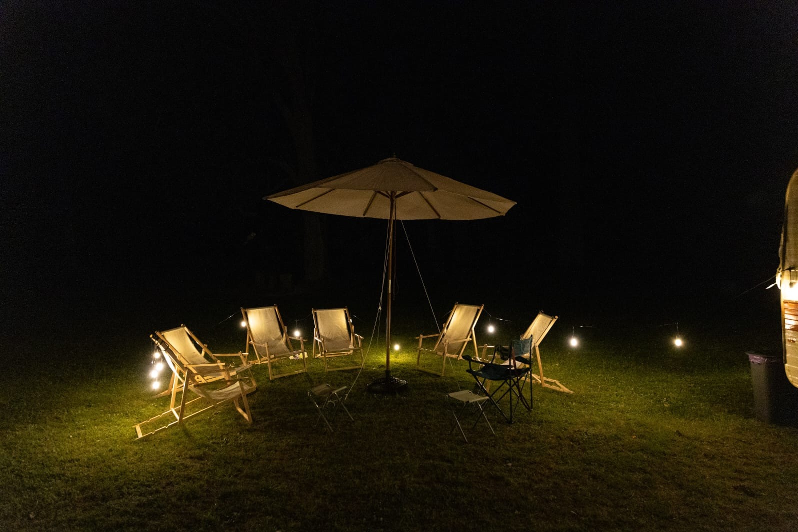 Nocna sceneria z parasolem, krzesłami plażowymi i girlandą świetlną na trawie, tworząca kameralną atmosferę. Przyczepa kempingowa w tle.