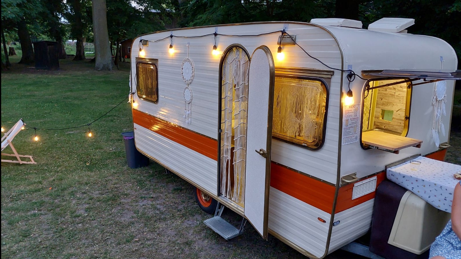 Oświetlona, kremowo-pomarańczowa fotobudka w stylu retro na zewnątrz, z otwartymi drzwiami i oknem serwującym, udekorowana łapaczami snów i girlandą żarówek.
