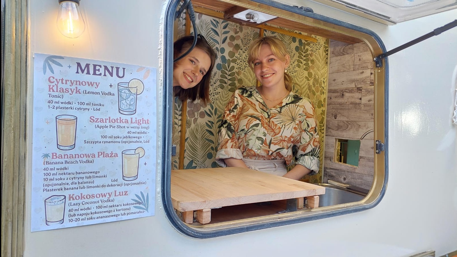 Uśmiechnięte barmanki w food trucku z menu drinków w Miechowej. Dekoracyjne tapety i drewniany blat baru tworzą przytulną atmosferę.