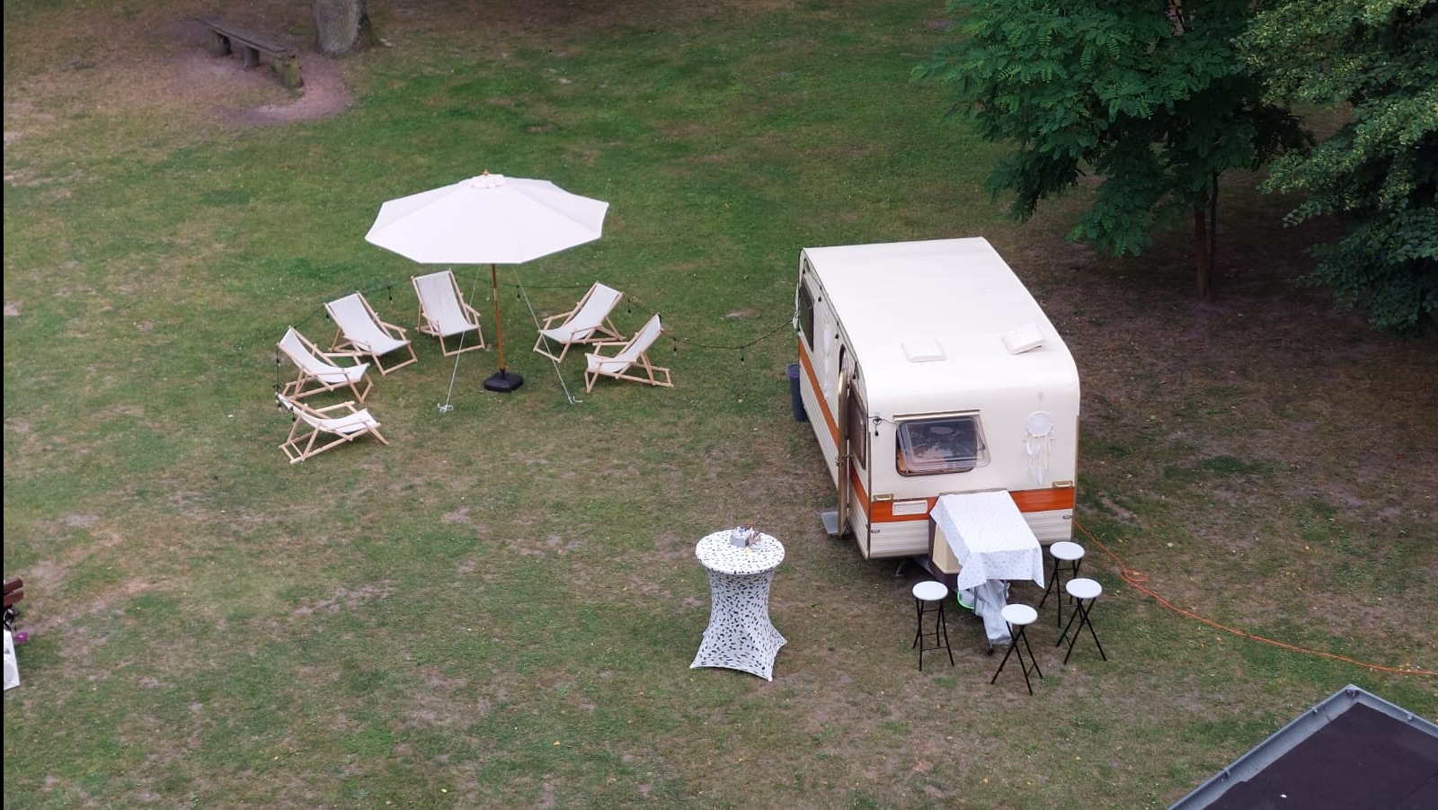 Widok z góry na przyczepę kempingową z dekoracjami, leżakami i stolikiem na trawie, sugerujący plenerową imprezę lub sesję zdjęciową w stylu vintage.