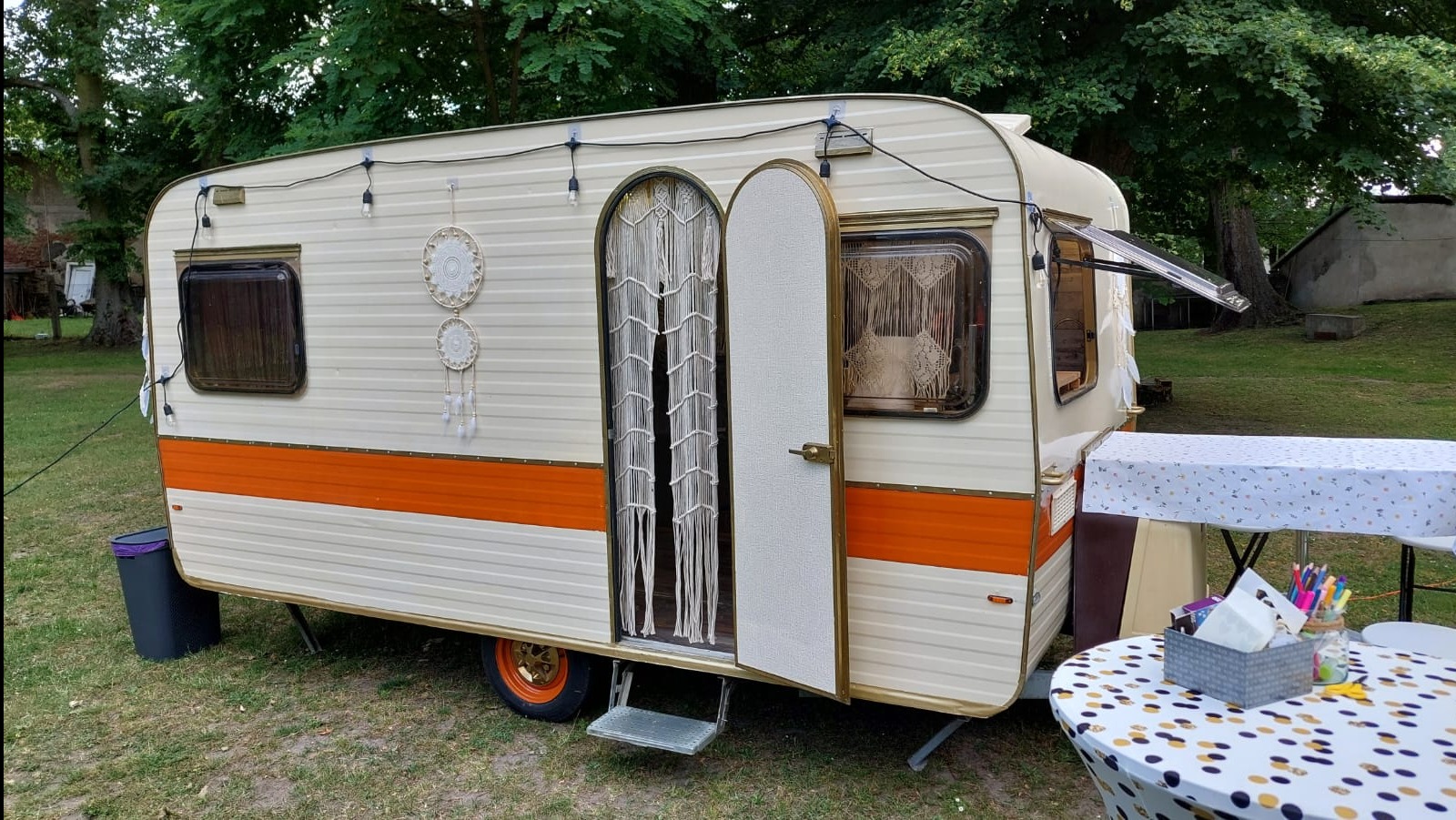 Klimatyczna fotobudka w stylu retro: kremowy kamper z pomarańczowym pasem, ozdobiony łapaczami snów i girlandą świetlną, otwarte drzwi z makramą, stoliki obok.