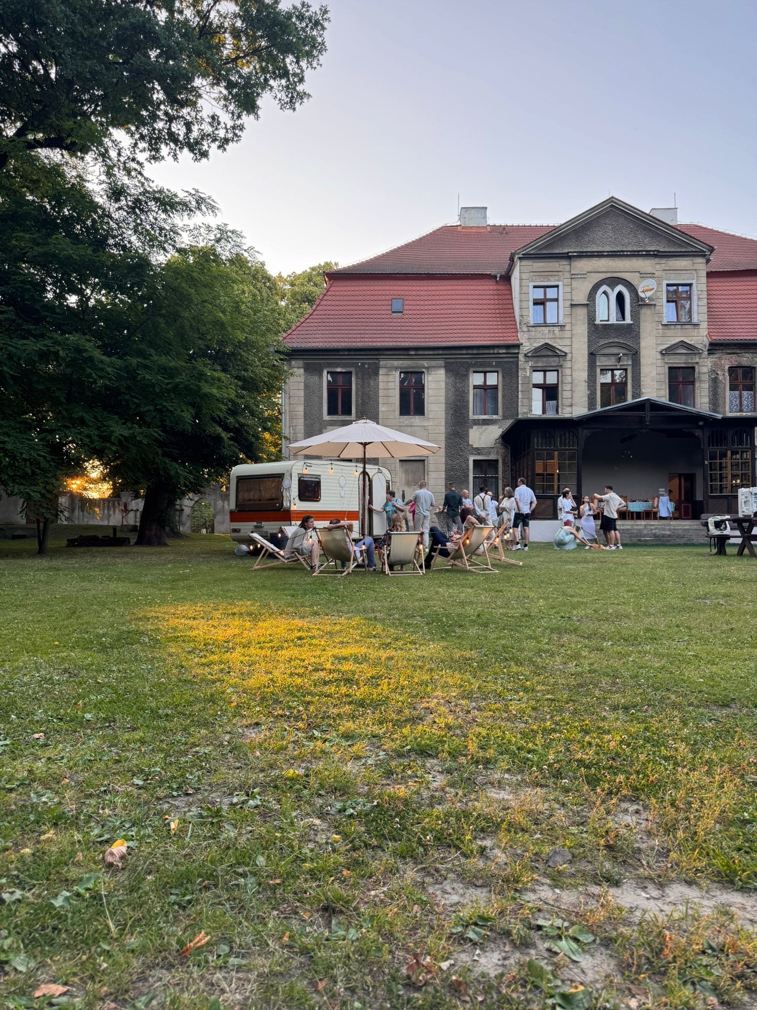 Plenerowa impreza z przyczepą kempingową i parasolem na tle starego budynku. Ludzie siedzą na leżakach i rozmawiają. Zachodzące słońce dodaje ciepła.