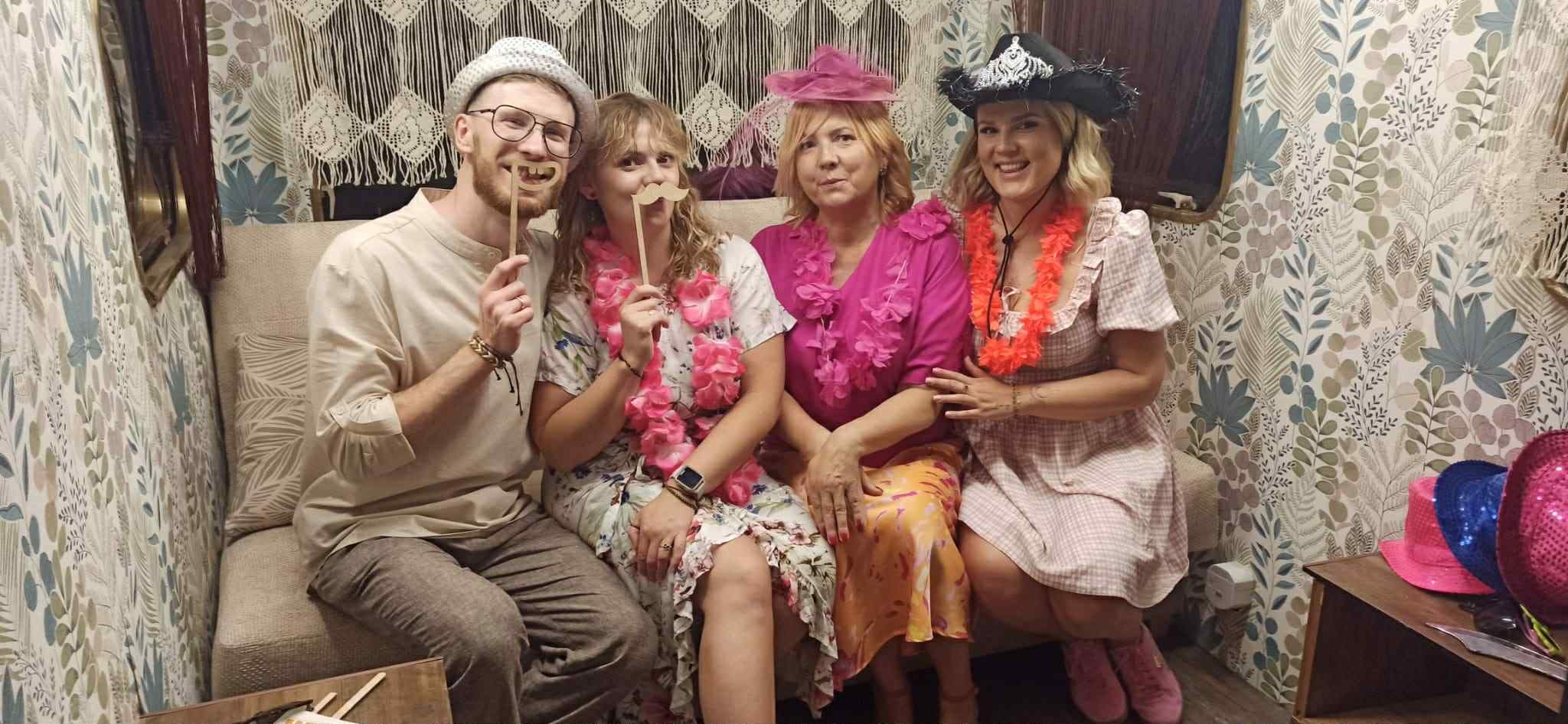 Uśmiechnięta grupa czterech osób w fotobudce, z rekwizytami (wąsy, zęby) i kwiatowymi girlandami, pozuje na tle wzorzystej tapety i dekoracji z makramy.