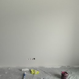 Nowik zlota raczka - Świeżo pomalowane na biało ściany pokoju, widoczne narzędzia malarskie i puszka farby. Otwór drzwiowy zabezpieczony taśmą malarską. Podłoga przykryta folią.