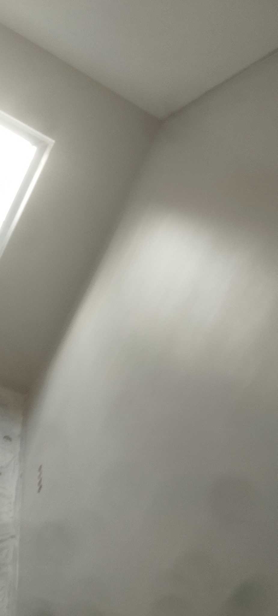 Świeżo pomalowana, gładka ściana w odcieniach szarości, z widocznym fragmentem okna w lewym górnym rogu. Minimalistyczne wykończenie wnętrza.