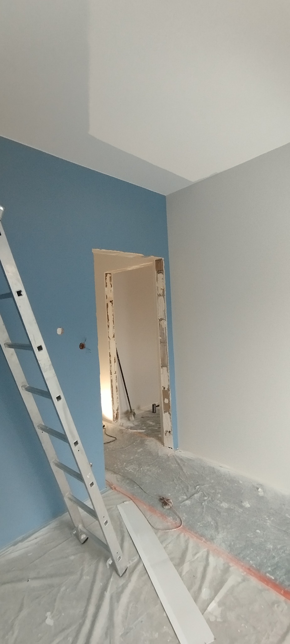 Pokój w trakcie malowania: ściana błękitna, druga biała. Widoczna drabina i zabezpieczona folią podłoga. Ościeżnica drzwiowa w surowym stanie.