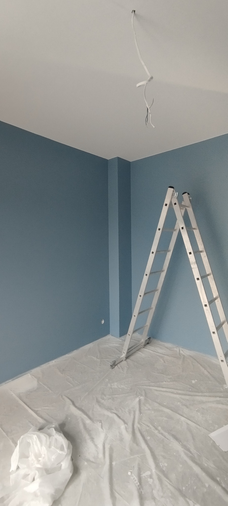 Ściana pomalowana na niebiesko, drabina aluminiowa oparta o ścianę, podłoga zabezpieczona folią malarską. Wystające przewody z sufitu.