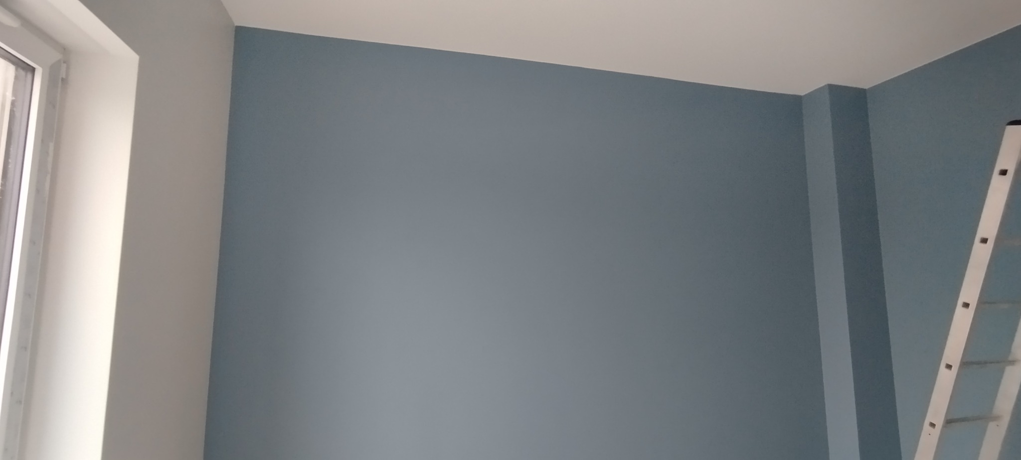 Świeżo pomalowane na szaro ściany z widoczną drabiną malarską i fragmentem okna.  Gładkie wykończenie i jasne, neutralne oświetlenie.
