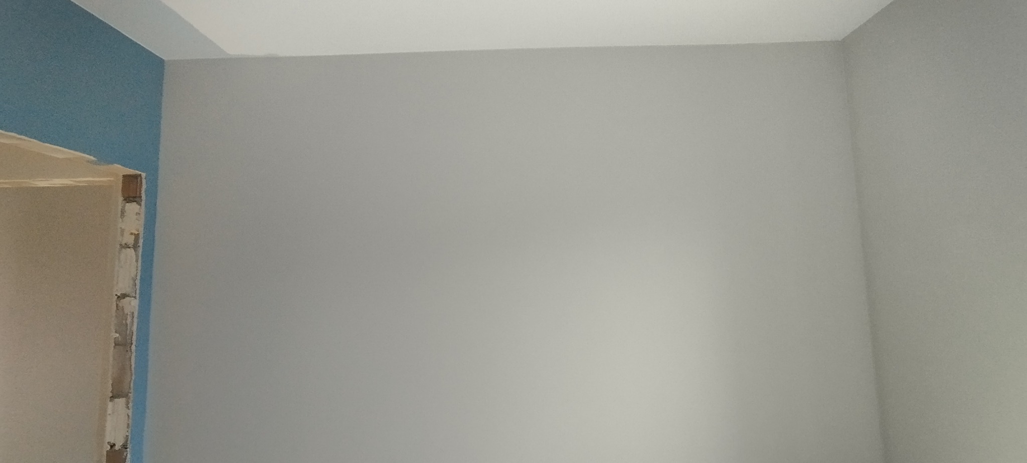 Pomieszczenie po malowaniu: szara ściana z niebieskim akcentem przy ościeżnicy. Gładkie powierzchnie, świeży wygląd. Wnętrze w trakcie wykańczania.