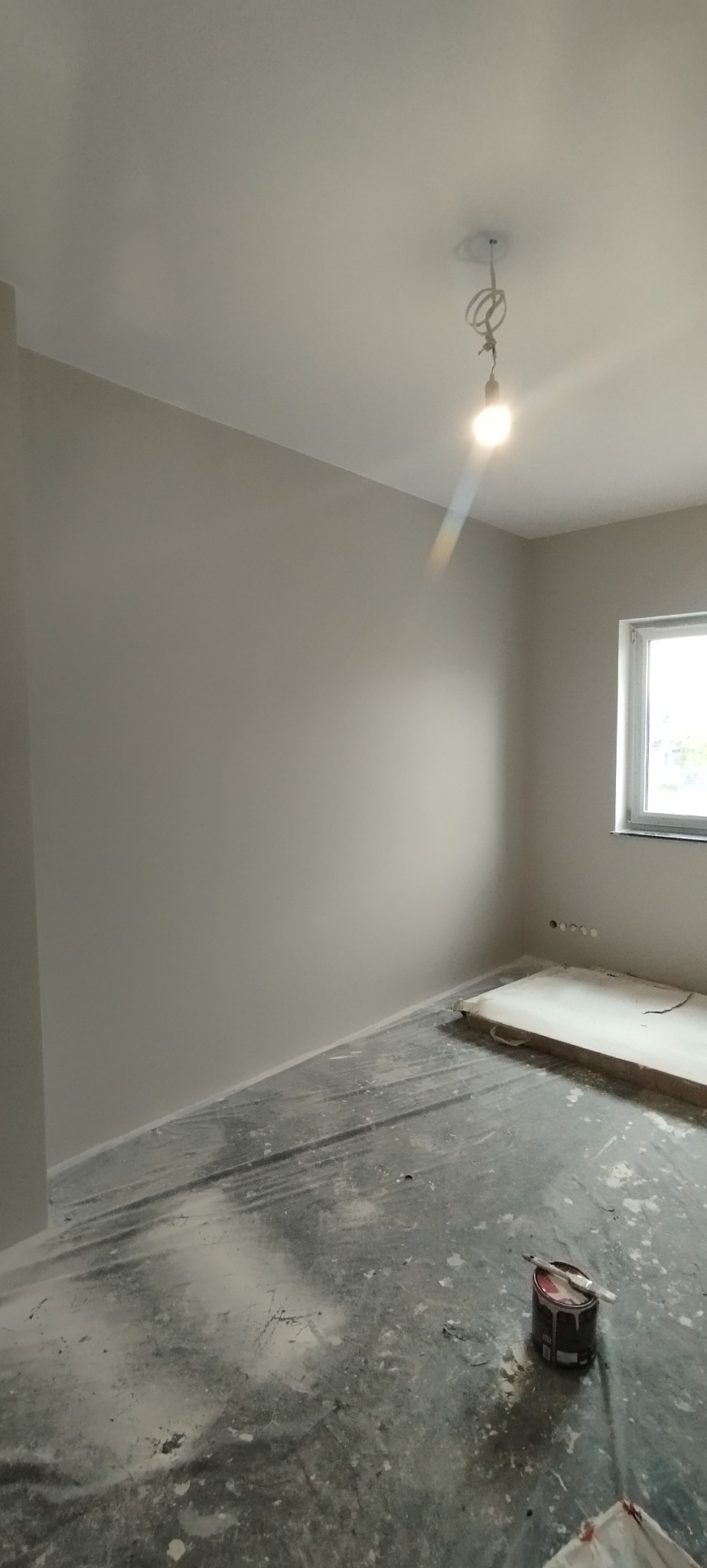 Pokój po malowaniu na szaro, z widoczną puszką farby i pędzlem na podłodze. Przy oknie widoczne gniazdka elektryczne. W tle surowy kabel z żarówką.