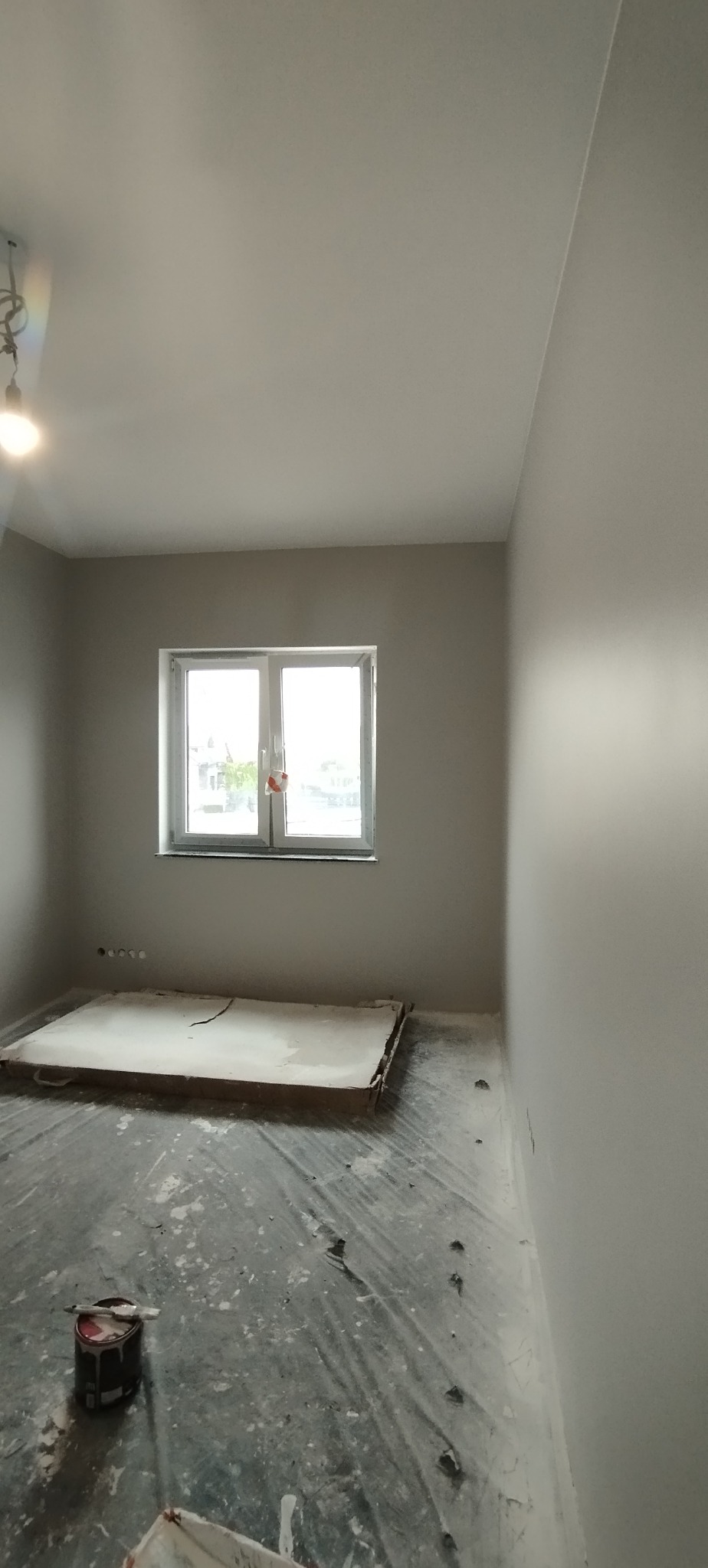 Wnętrze po malowaniu ścian na szaro, widoczne okno, zabezpieczona podłoga folią i puszka z farbą. Świeżo pomalowane ściany w pokoju.