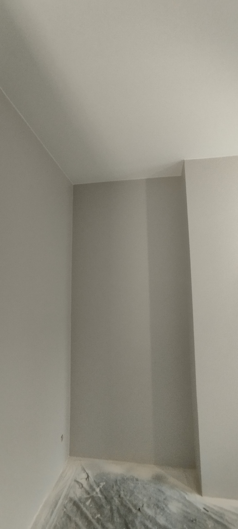 Jasne, świeżo pomalowane ściany w rogu pokoju, z widocznym zabezpieczeniem podłogi folią malarską. Minimalistyczne wykończenie wnętrza w neutralnej kolorystyce.