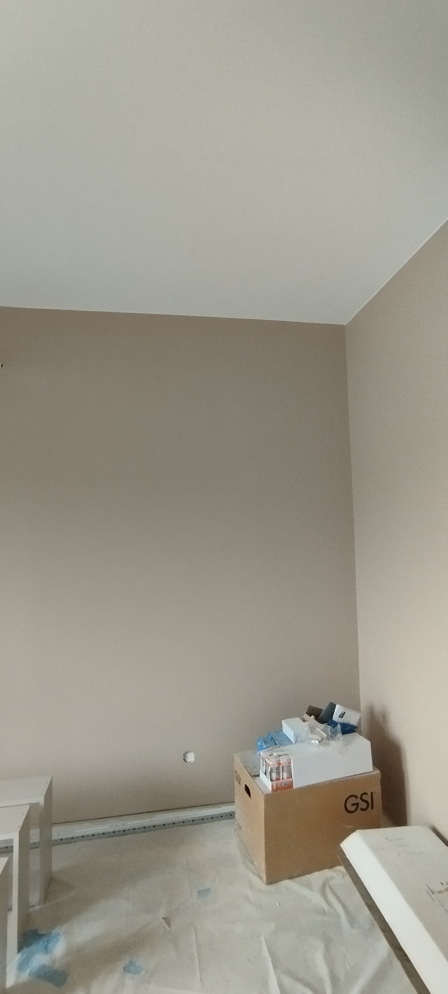 Ściana po malowaniu na beżowo, zabezpieczona podłoga, karton z akcesoriami. Widok na pomalowane wnętrze w trakcie prac wykończeniowych.