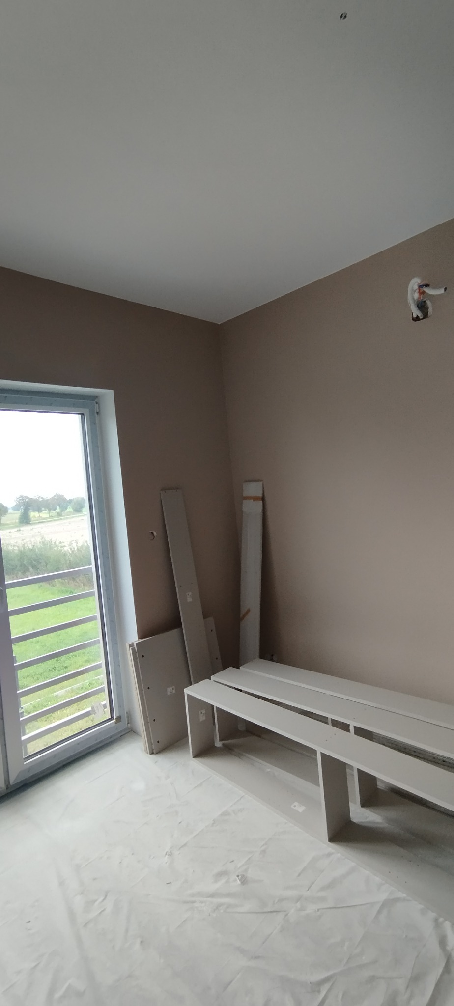 Pokój po malowaniu ścian na beżowo, z białym sufitem i zabezpieczoną folią podłogą. Widoczne płyty karton-gips i listwy przypodłogowe przy oknie i ścianie.