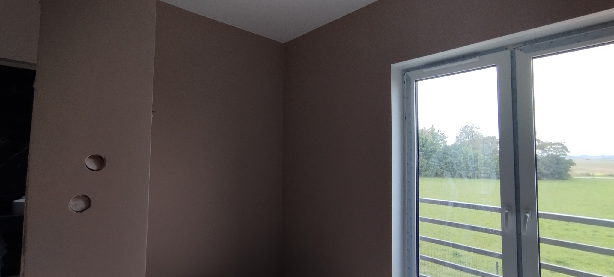 Ściana w kolorze beżowym z widocznymi otworami na gniazdka elektryczne i białym oknem z widokiem na zieleń. Minimalistyczny styl wnętrza.