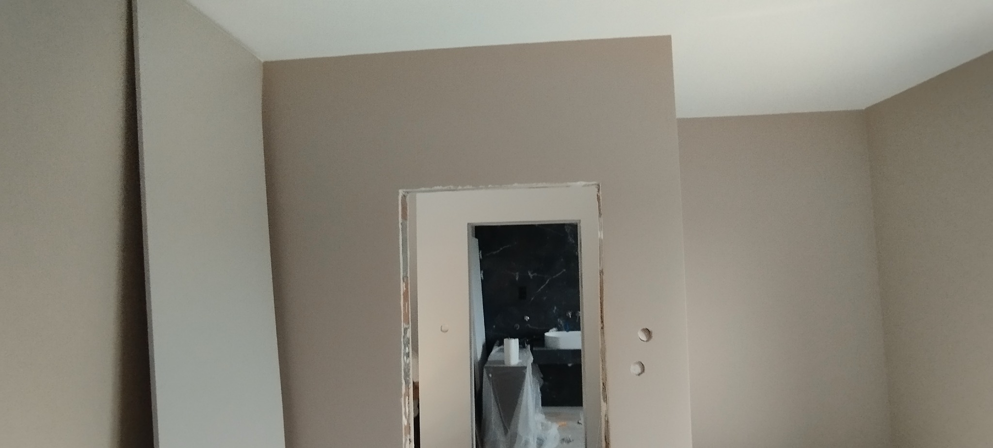 Wnętrze w trakcie remontu: świeżo pomalowane ściany w beżowym odcieniu, widoczny otwór drzwiowy z wykończoną ościeżnicą, w tle wykończona łazienka z czarnym marmurem.