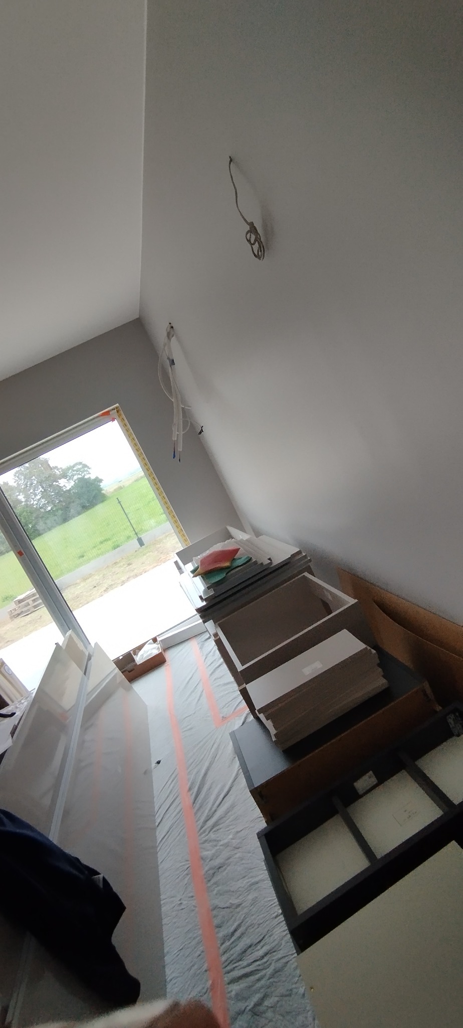 Poddasze z oknem: przygotowane elementy mebli, zabezpieczona podłoga folią malarską, kable zwisające z sufitu. Etap prac wykończeniowych przed montażem.