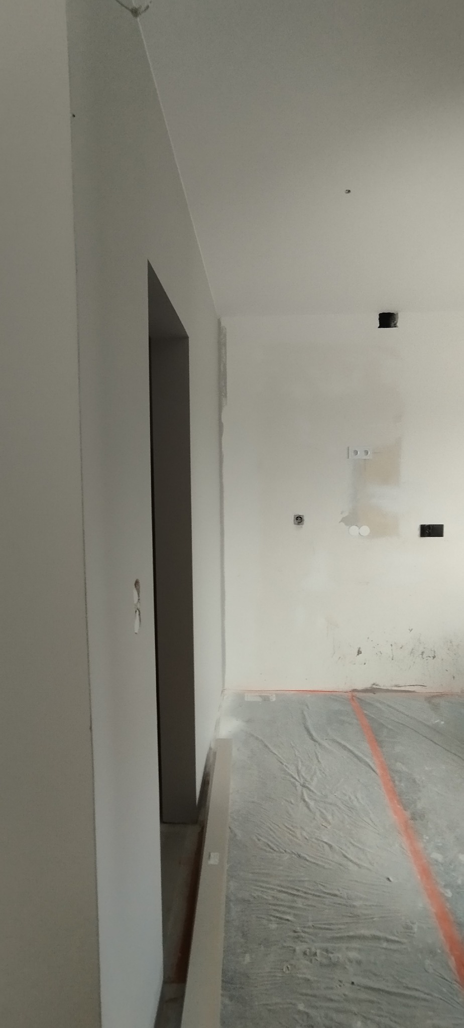 Wnętrze w trakcie remontu: świeżo pomalowane ściany na biało, zabezpieczona podłoga folią malarską, widoczne puszki elektryczne i otwór drzwiowy.