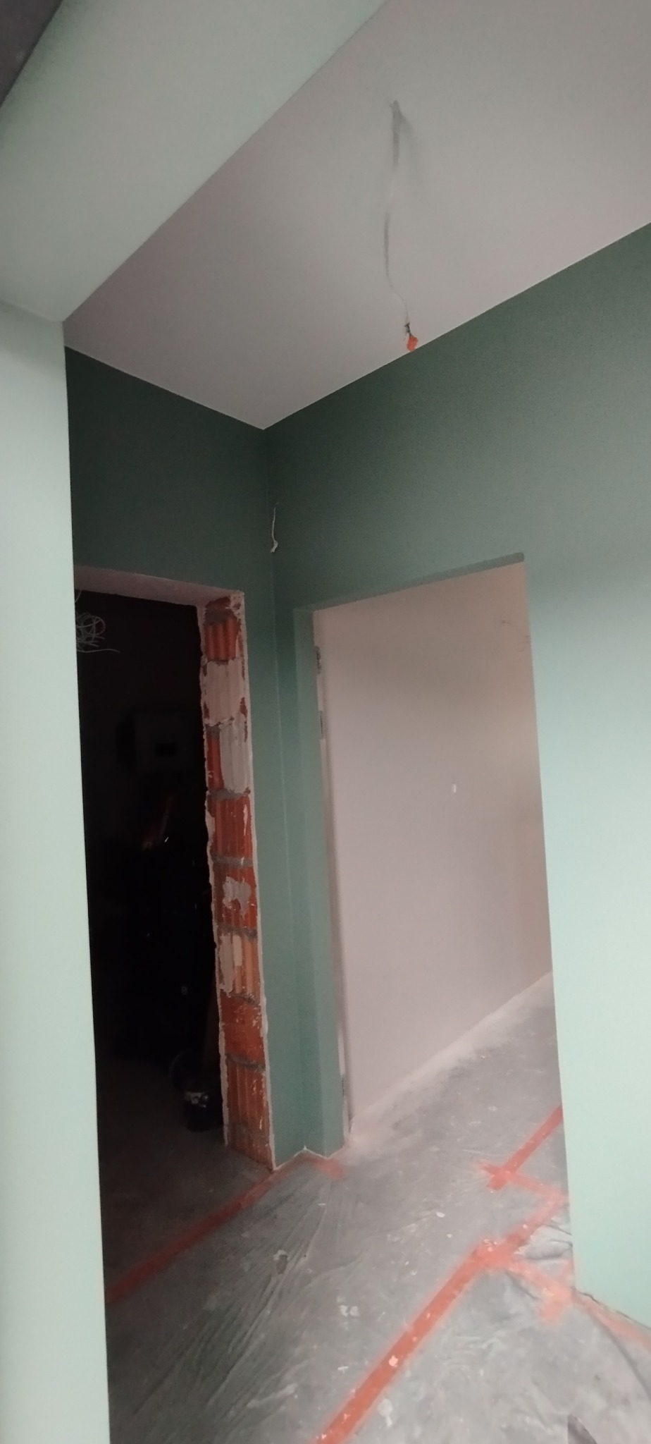 Wnętrze w trakcie odświeżania: pomalowane ściany na zielono, surowa cegła wokół otworu drzwiowego, zabezpieczona folią podłoga, widoczne przewody elektryczne.