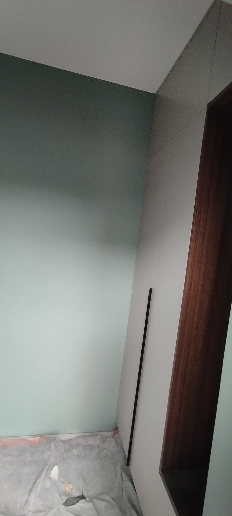 Świeżo pomalowane na jasnozielono ściany w rogu pokoju, z widoczną częścią białej szafy z drewnianymi elementami i zabezpieczoną folią podłogą.