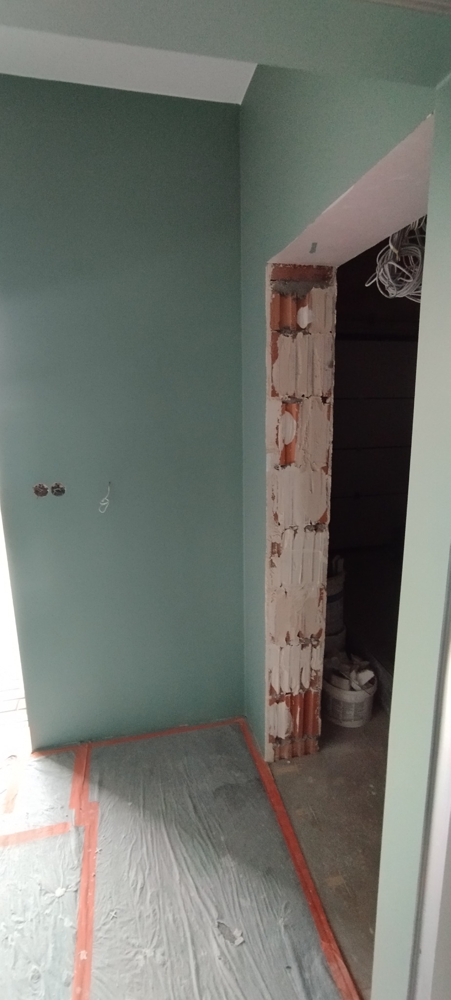 Ściana w kolorze miętowym po malowaniu, zabezpieczona podłoga folią, ościeżnica z cegły w trakcie obróbki. Widoczne kable. Prace wykończeniowe w pomieszczeniu.