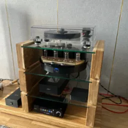 Drewniany regał audio z półkami ze szkła, na którym umieszczono gramofon i wzmacniacz lampowy. Widoczne kable i elementy audiofilskiego sprzętu.