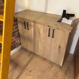 Paweł Kowalik - Drewniana szafka z czarnymi uchwytami, obok drabina i drzwi. Na blacie akcesoria montażowe i pudełka. Podłoga z paneli. Widok na szafkę w pomieszczeniu.