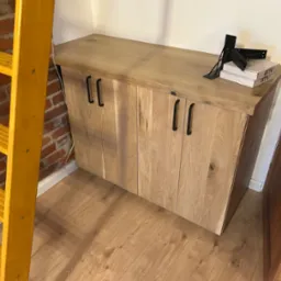 Drewniana szafka z czarnymi uchwytami, obok drabina i drzwi. Na blacie akcesoria montażowe i pudełka. Podłoga z paneli. Widok na szafkę w pomieszczeniu.