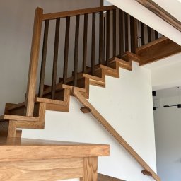 Paweł Kowalik - Jasne drewniane schody z ciemnymi balustradami na białej ścianie. Widok z dołu, ukazujący solidną konstrukcję i nowoczesny design.