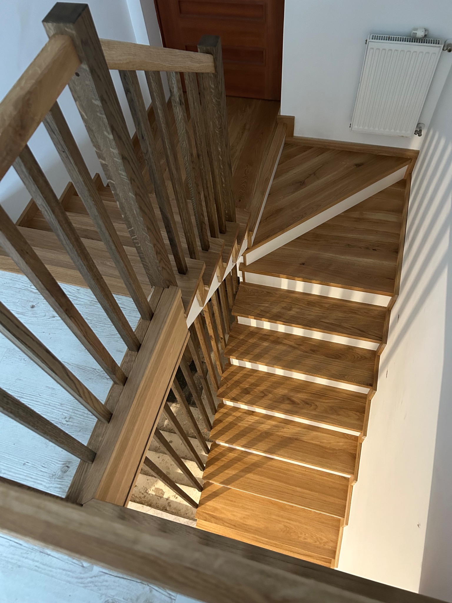 Drewniane schody z balustradą, widok z góry. Stopnie w odcieniach beżu, białe podstopnice. U góry widoczny fragment drewnianych drzwi i grzejnika.