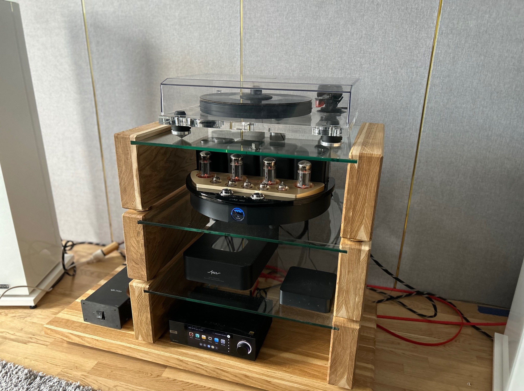 Drewniany regał audio z półkami ze szkła, na którym umieszczono gramofon i wzmacniacz lampowy. Widoczne kable i elementy audiofilskiego sprzętu.
