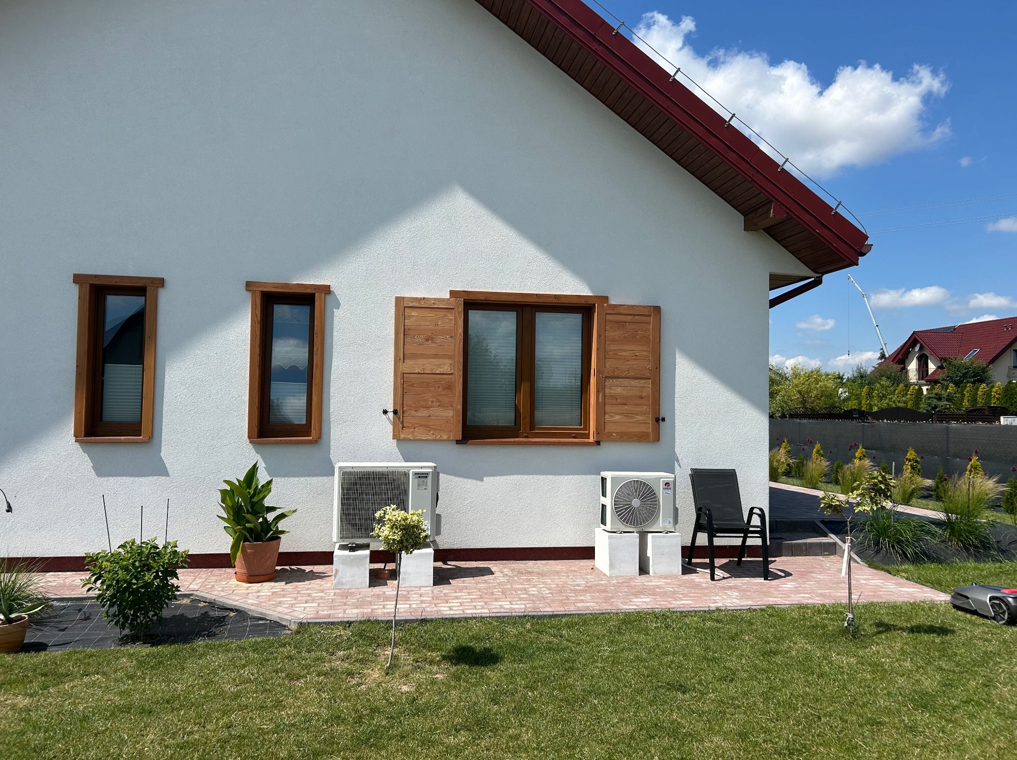 Biały dom z drewnianymi okiennicami i ramami okiennymi. Widok na ogród z roślinami w donicach i robotem koszącym. Jasne, słoneczne oświetlenie.