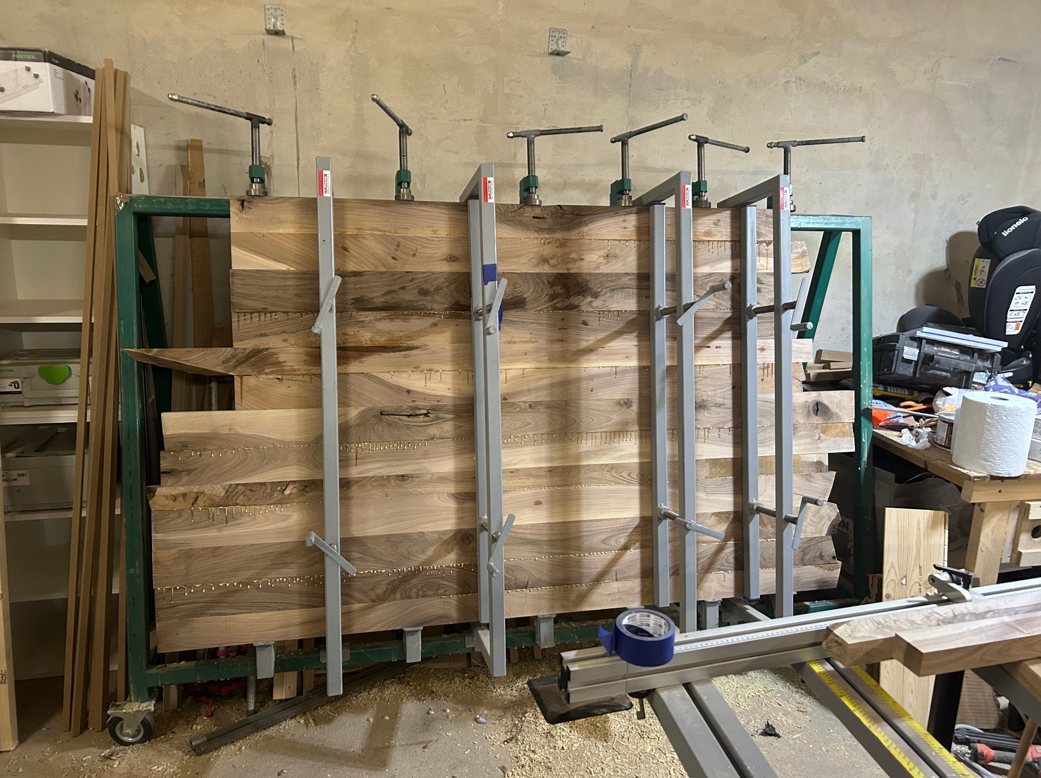 Sklejanie desek z drewna orzechowego za pomocą ścisków stolarskich. Widoczne krople kleju. Deski umieszczone w zielonej ramie. W tle półki i narzędzia.