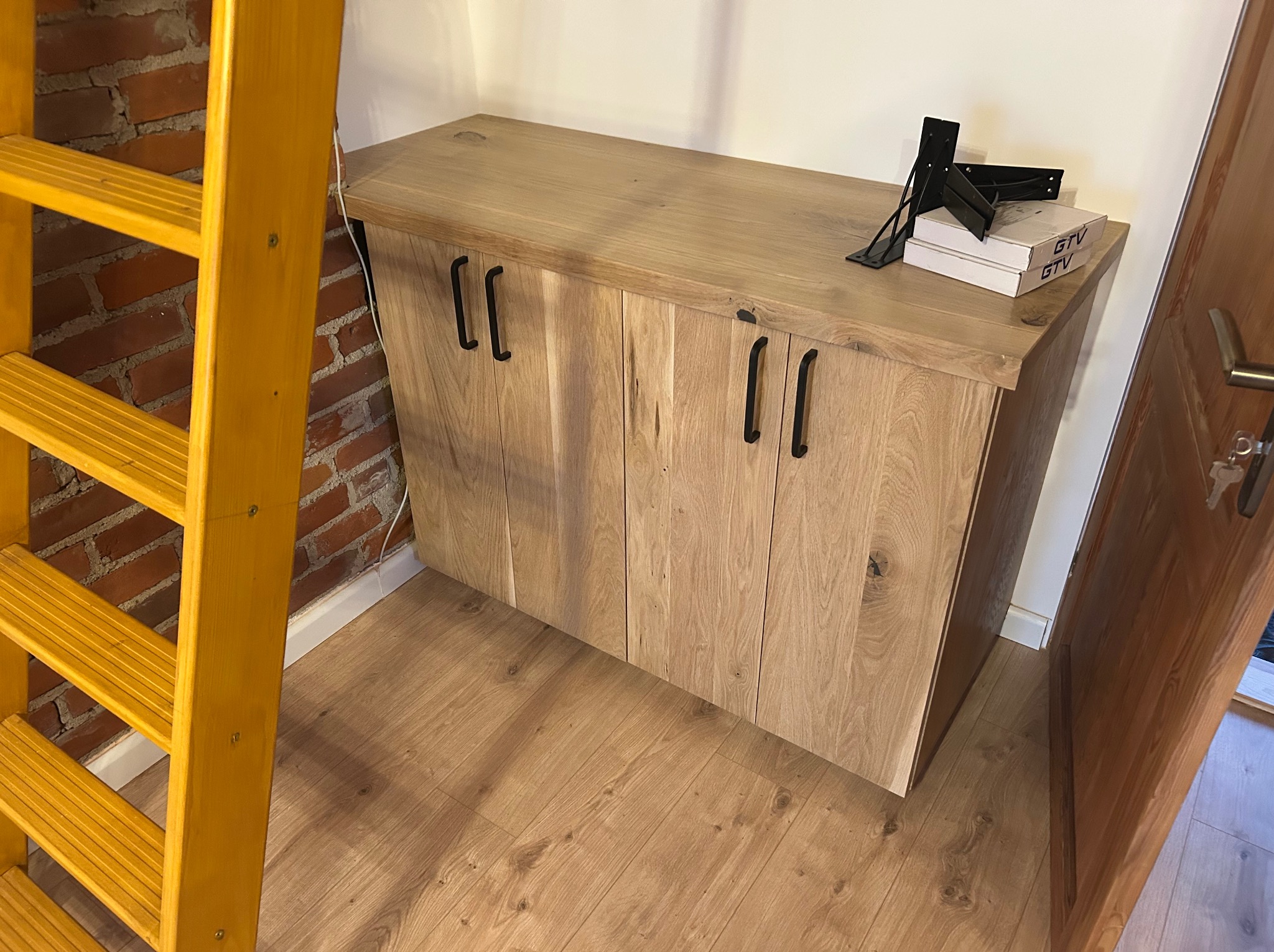 Drewniana szafka z czarnymi uchwytami, obok drabina i drzwi. Na blacie akcesoria montażowe i pudełka. Podłoga z paneli. Widok na szafkę w pomieszczeniu.