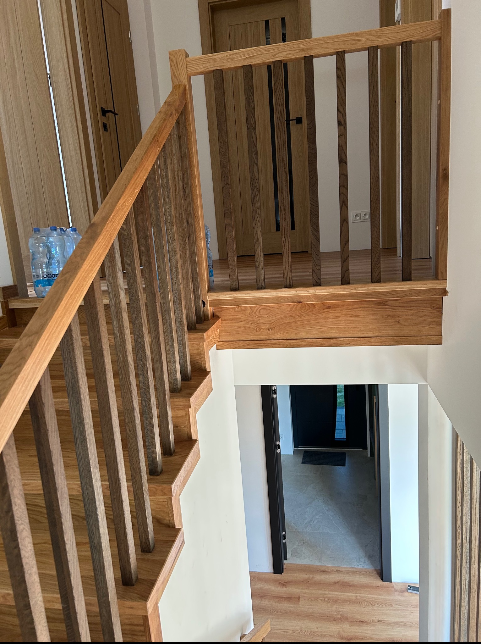 Jasne, drewniane schody z balustradą, widoczne z góry, z butelkami wody na podeście. W tle drzwi i fragment podłogi.