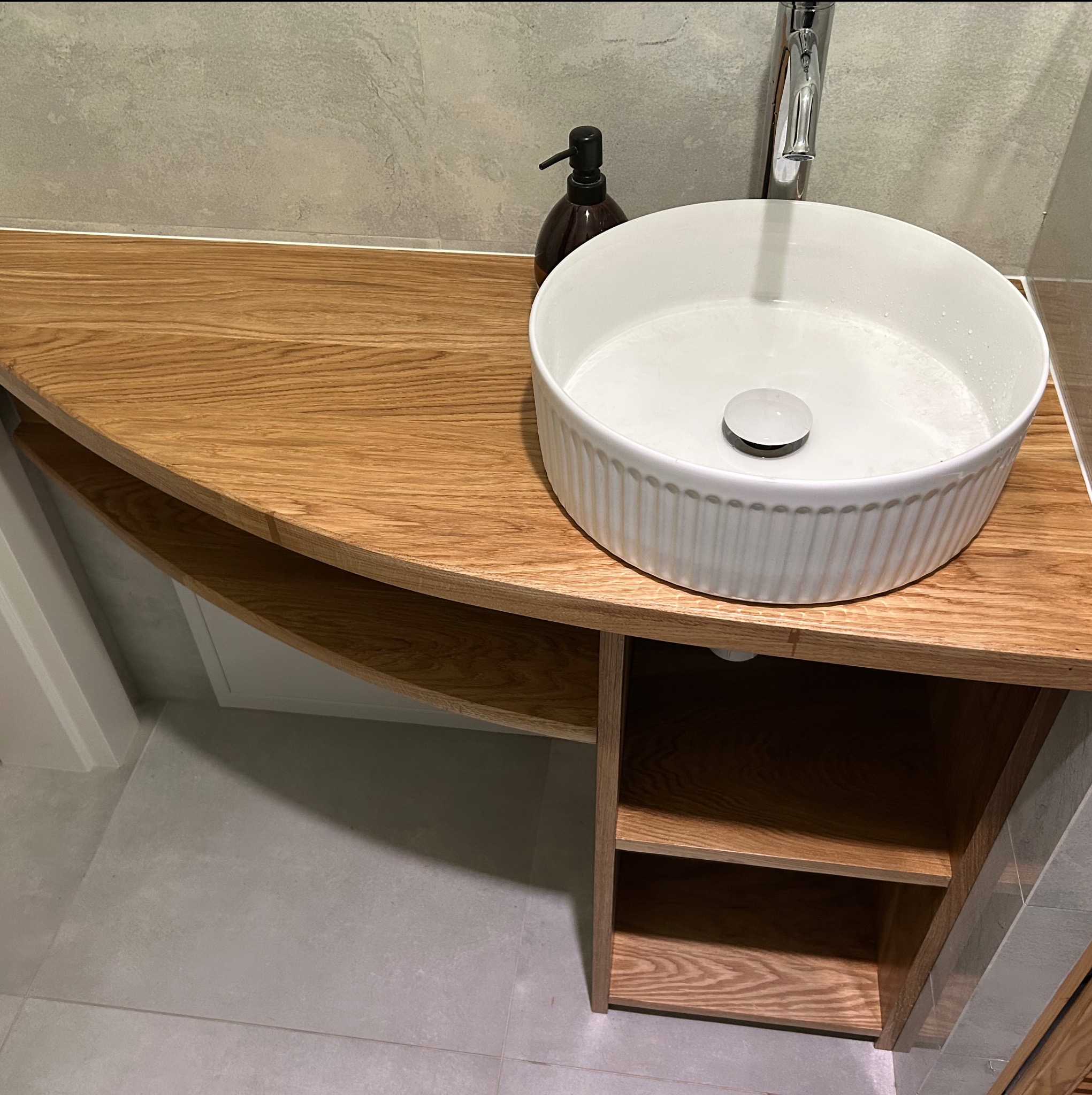 Okrągła umywalka na blacie z drewna dębowego z półkami, minimalistyczna łazienka w szarościach. Widoczny dozownik mydła i chromowana bateria.