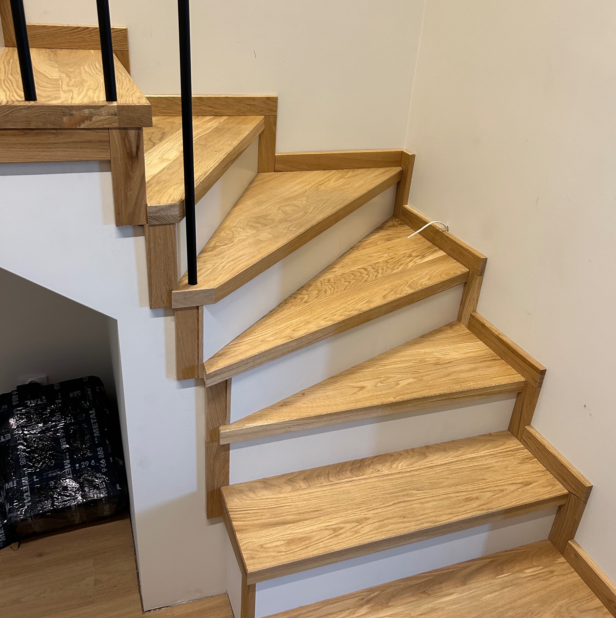 Drewniane schody z jasnego drewna, białe podstopnice, czarne barierki. Widoczne detale wykończenia i pakunek folii na podłodze.