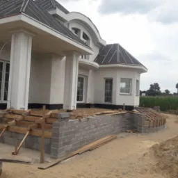 Budowa domu w stylu dworkowym: wzniesione fundamenty schodów wejściowych z szarego betonu komórkowego, częściowo oszalowane drewnem. Widok na elewację i obrzeża budynku.