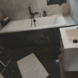 ZAKŁAD OGOLNOBUDOWLANY - Nowoczesna łazienka po remoncie: biała wanna z czarną armaturą, umywalka z szafką, płytki imitujące marmur na podłodze i ścianach. Widoczny mop i zabezpieczenie podłogi.