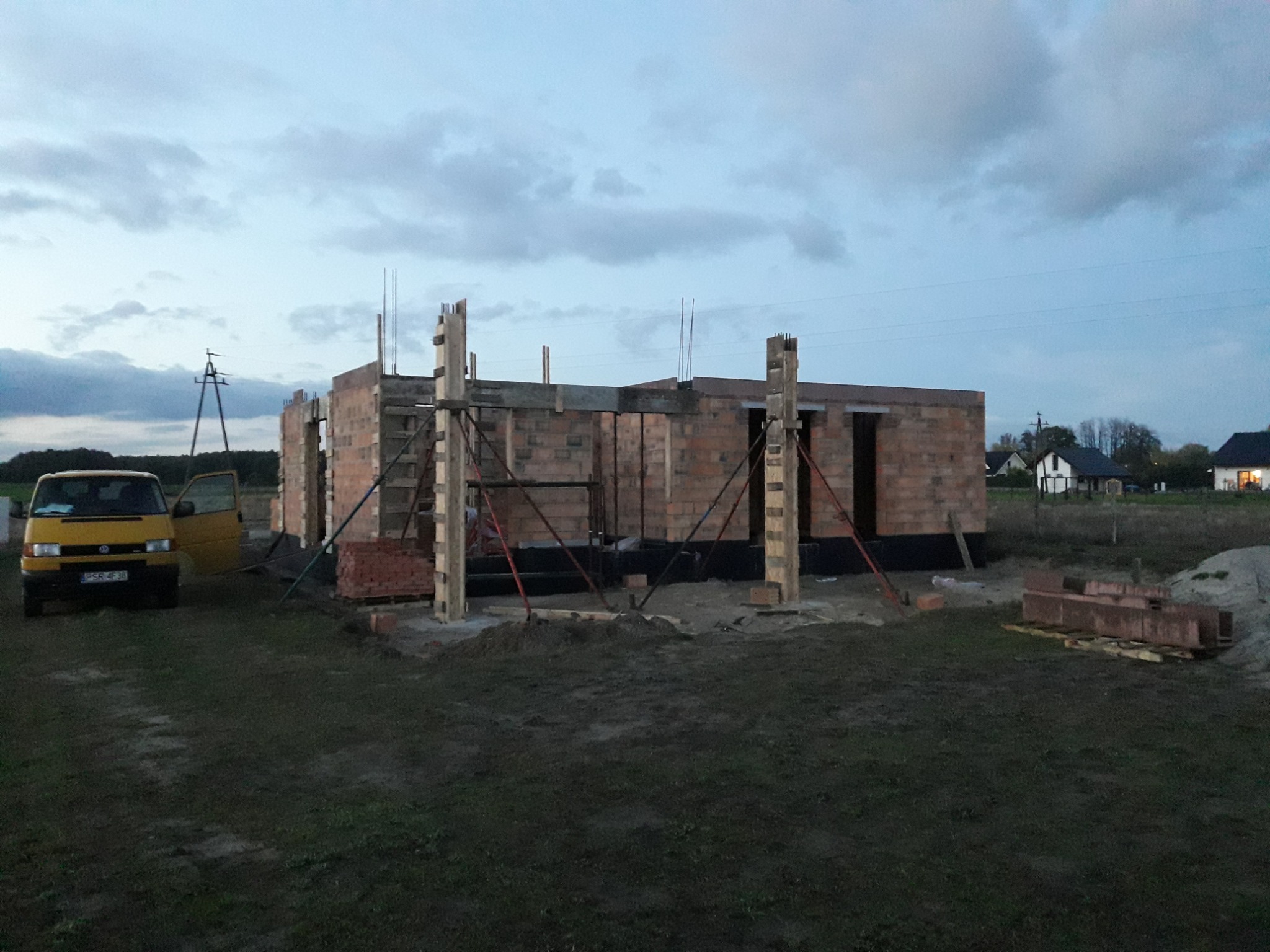 Wznoszenie ścian parterowego domu z czerwonej cegły, widoczne zbrojenia i szalunki kolumn. Żółty bus z otwartymi drzwiami stoi obok. Trawa i pochmurne niebo w tle.