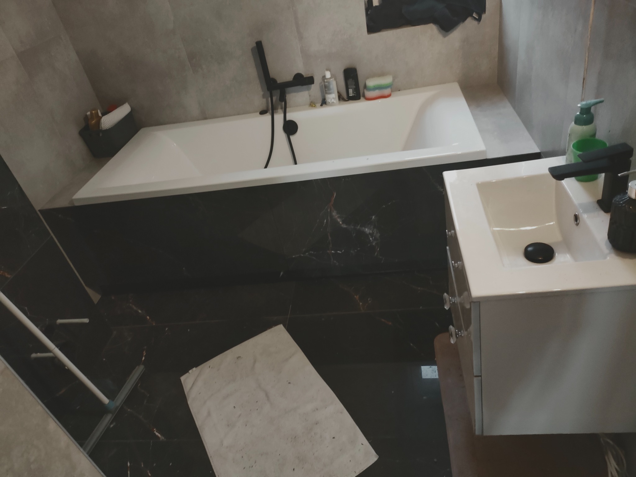 Nowoczesna łazienka po remoncie: biała wanna z czarną armaturą, umywalka z szafką, płytki imitujące marmur na podłodze i ścianach. Widoczny mop i zabezpieczenie podłogi.