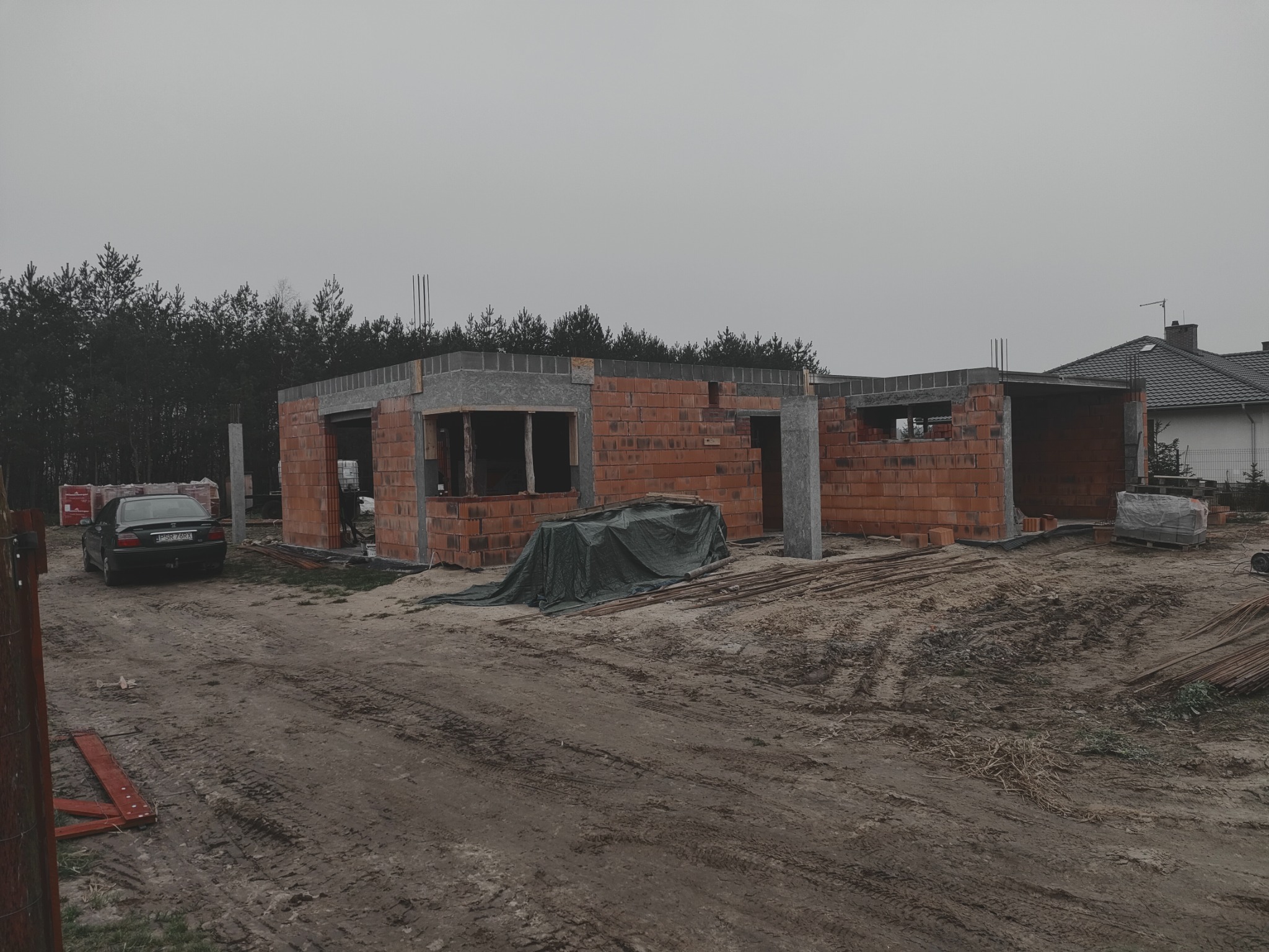 Budowa domu jednorodzinnego w stanie surowym otwartym: ceglane ściany, betonowe nadproża, zbrojenia. Na pierwszym planie teren budowy i zaparkowany samochód.