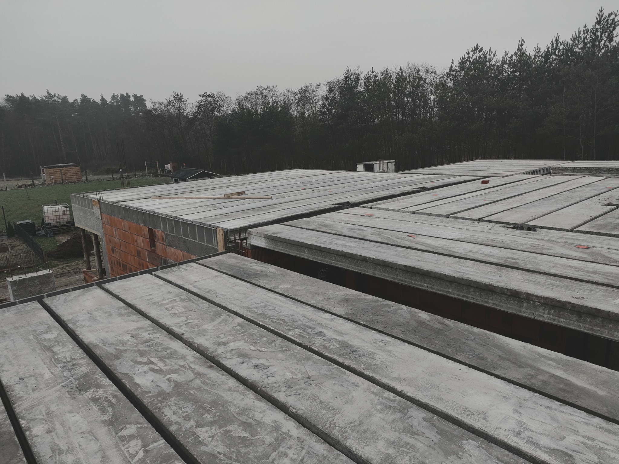 Budowa domu: strop Teriva z betonowych belek i pustaków, widok z góry na szary beton i czerwoną cegłę, las w tle, pochmurny dzień.