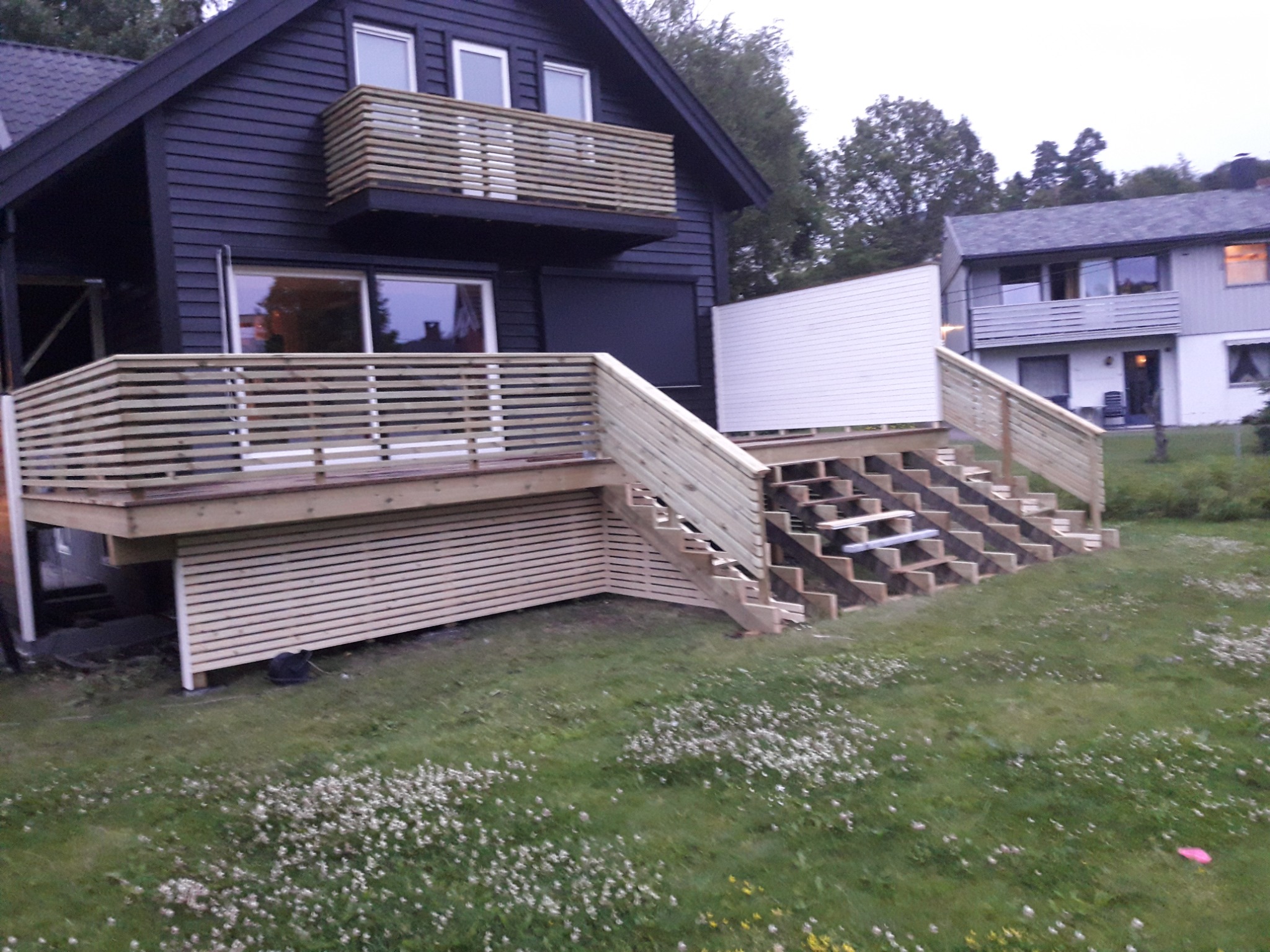 Nowa, drewniana konstrukcja tarasu z schodami przy ciemnym domu. Widoczne świeże deski i wykończenia. Drugi budynek w tle, trawa z kwiatami na pierwszym planie.