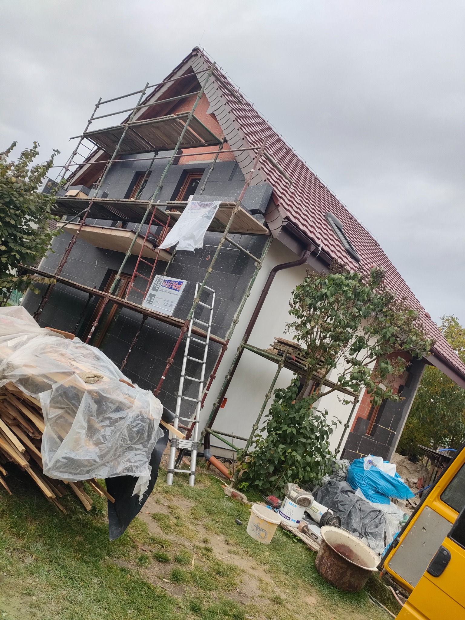 Dom jednorodzinny w trakcie ocieplania elewacji, widoczne rusztowania, płyty izolacyjne, okna z drewnianymi ramami i częściowo otynkowana ściana. Prace w toku.