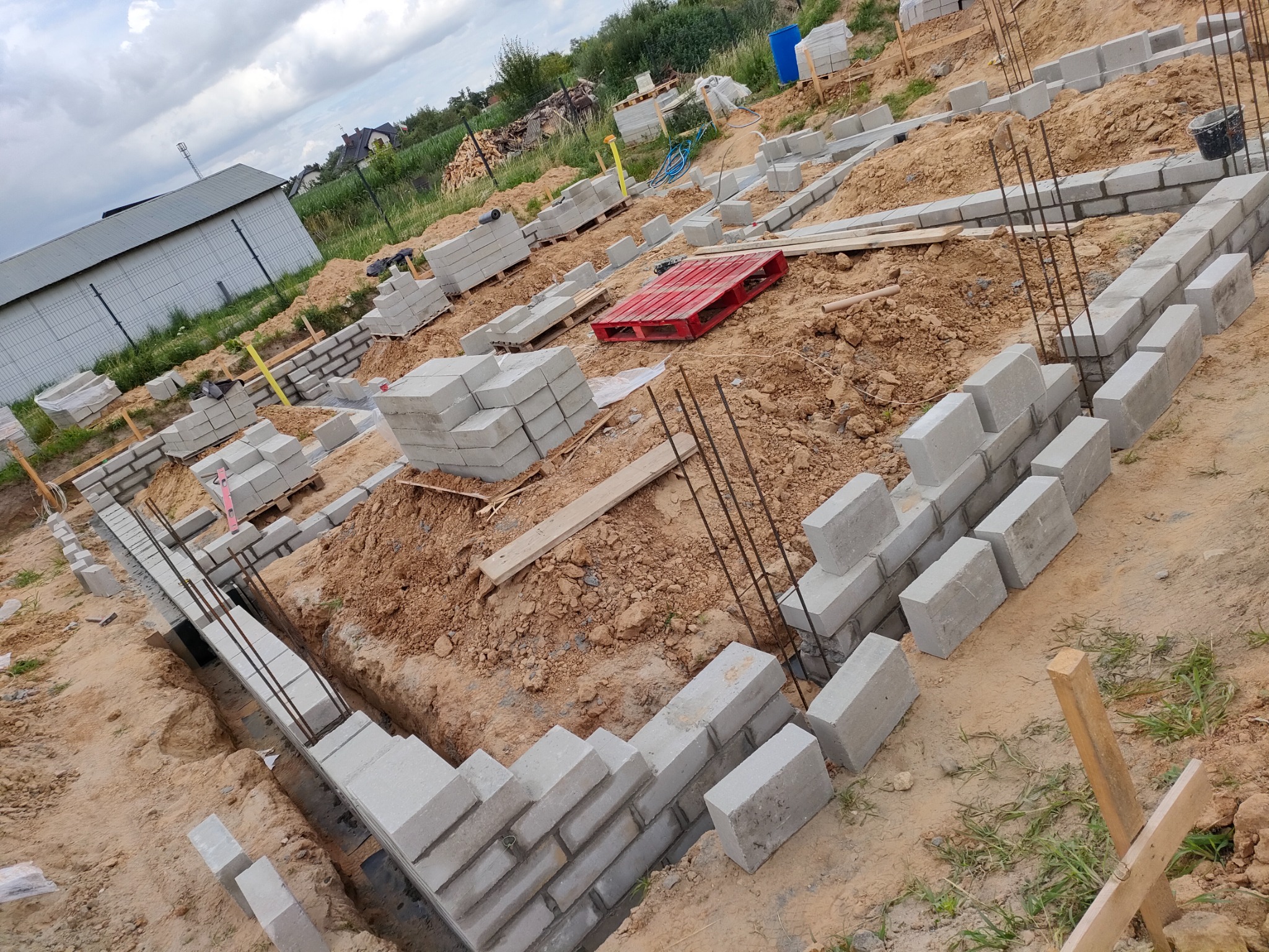 Wykop fundamentów z częściowo wymurowanymi ścianami z bloczków betonowych i wystającymi prętami zbrojeniowymi. Widoczny teren budowy z ziemią i materiałami budowlanymi.