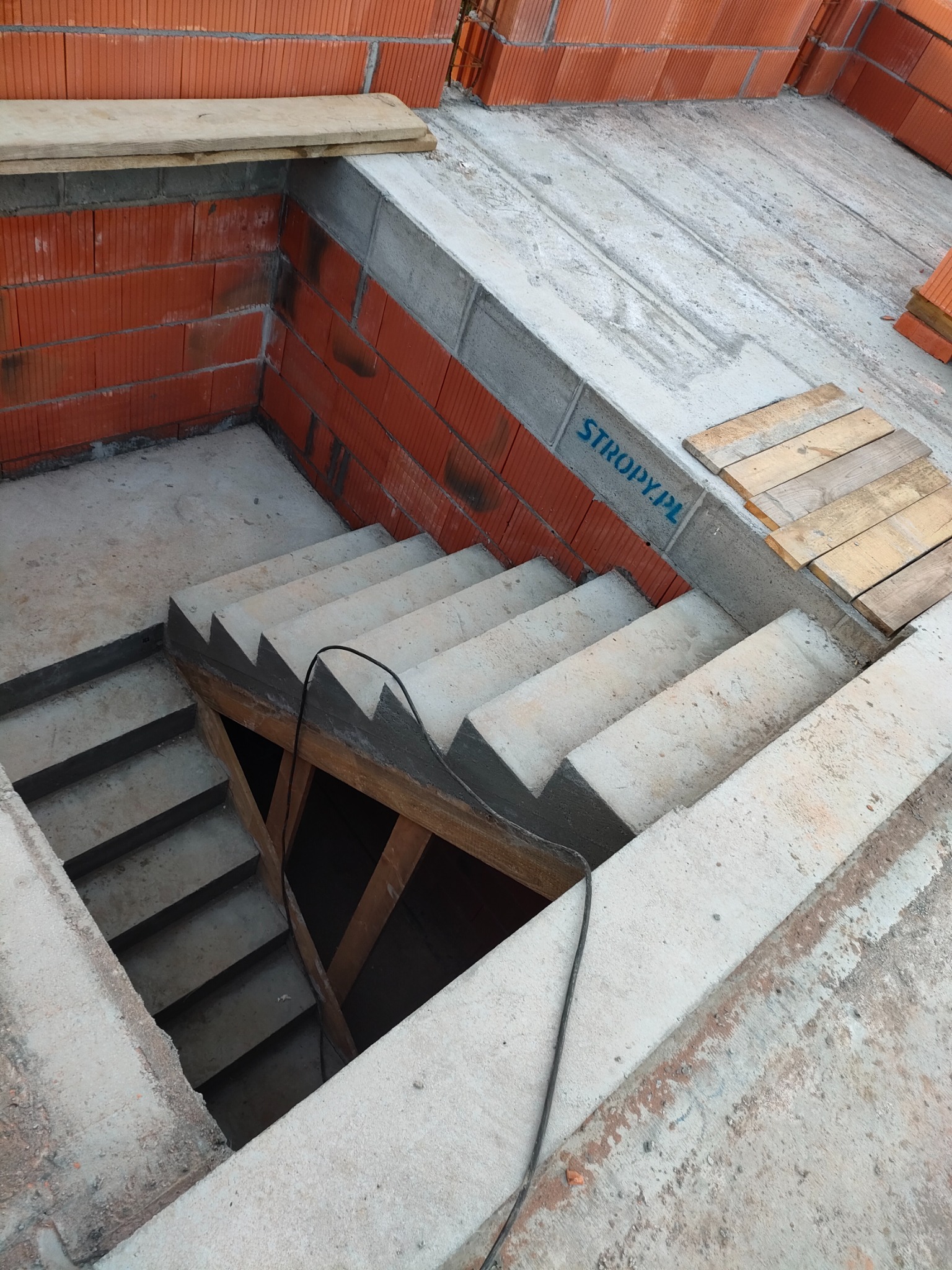 Surowe betonowe schody w dół, w trakcie budowy domu z czerwonej cegły. Widoczne drewniane podpory i fragment stropu z napisem 'STROPY.PL'.