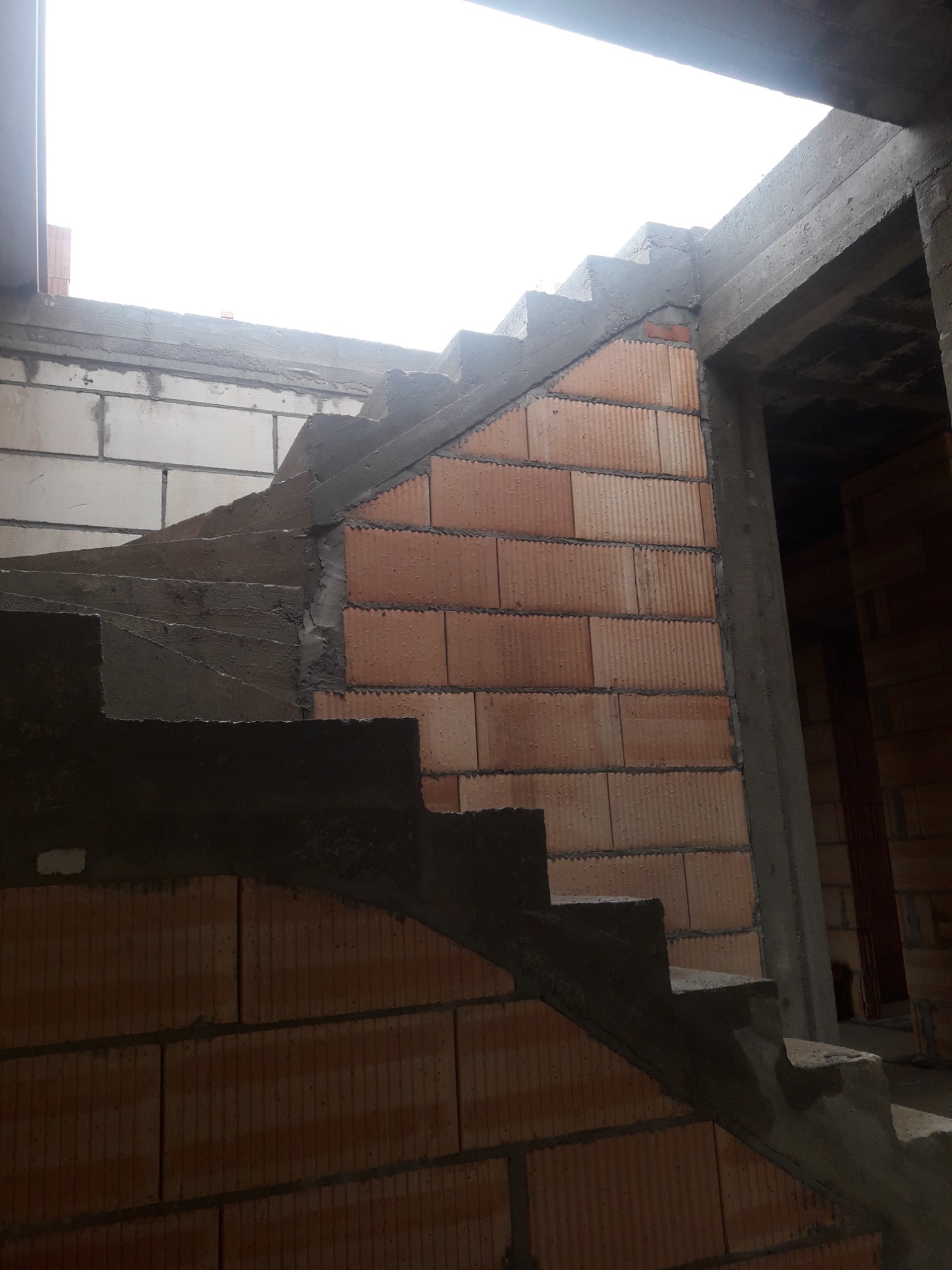 Wnętrze w trakcie budowy: spiralne schody z cegły i betonu, widoczne bloki z betonu komórkowego, surowy stan, brak wykończenia, oświetlenie górne.