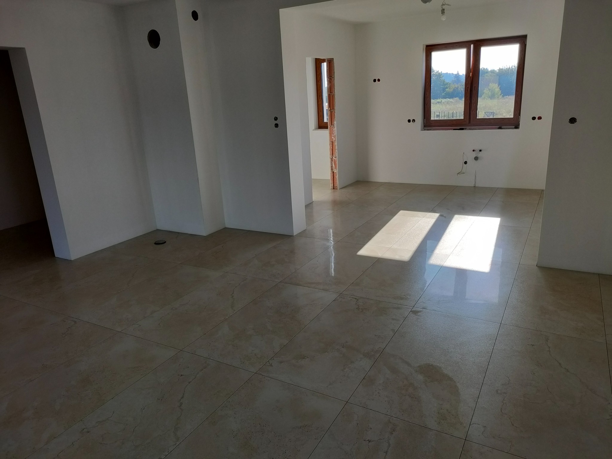 Jasne płytki podłogowe w nowym, przestronnym wnętrzu. Widoczne okna z drewnianą ramą i przygotowane instalacje elektryczne. Świeże, białe ściany.