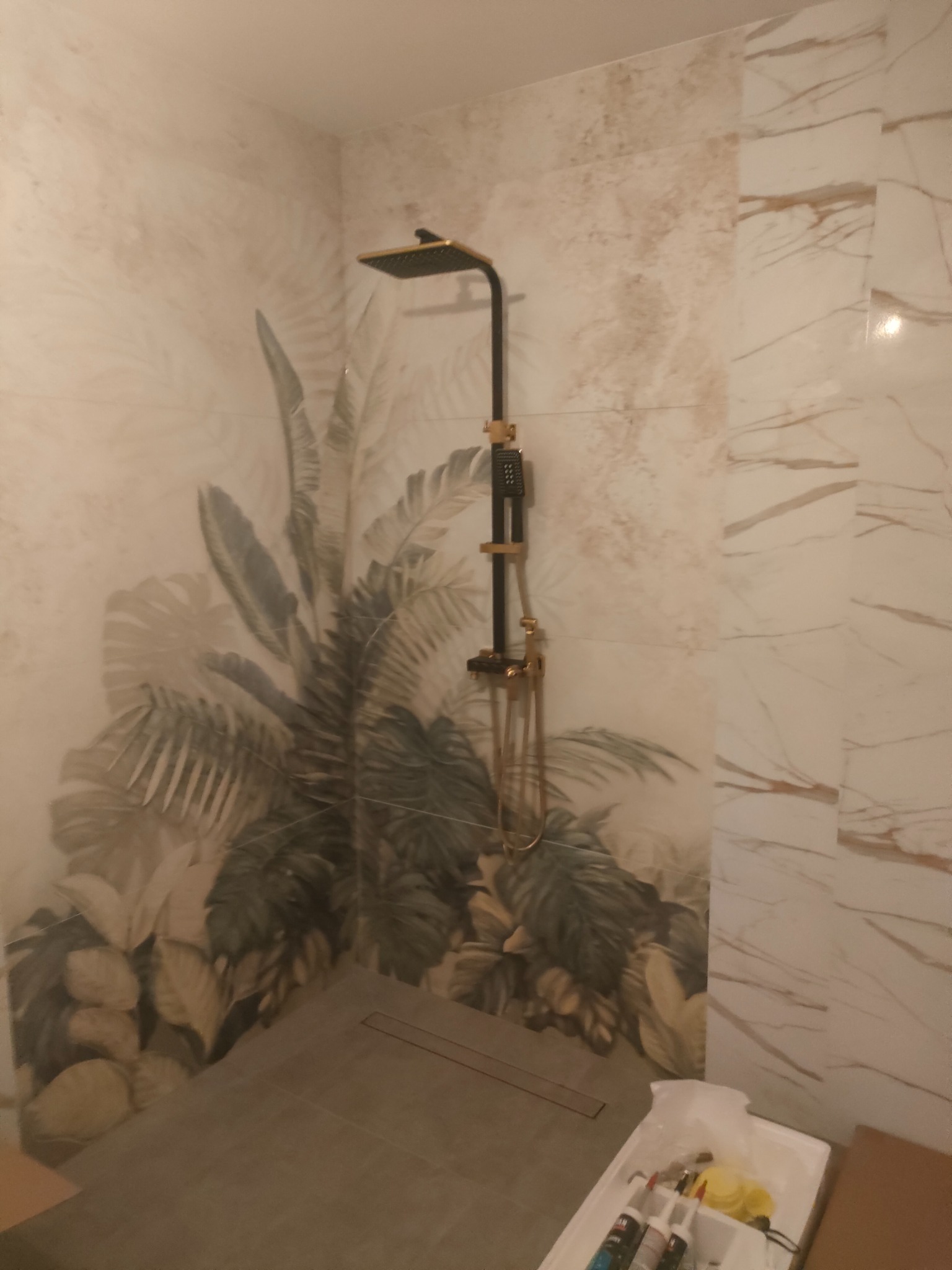 Nowoczesna kabina prysznicowa z deszczownicą w kolorze czarnym i złotym, wykończona płytkami z motywem roślinnym i marmurkowym. Widoczny odpływ liniowy.